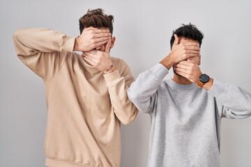 Young homosexual couple standing over white background covering eyes and mouth with hands, surprised and shocked. hiding emotion