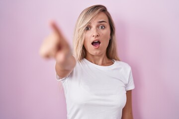 Poster - Young blonde woman standing over pink background pointing with finger surprised ahead, open mouth amazed expression, something on the front