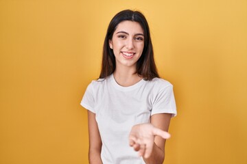 Young beautiful woman standing over yellow background smiling cheerful offering palm hand giving assistance and acceptance.