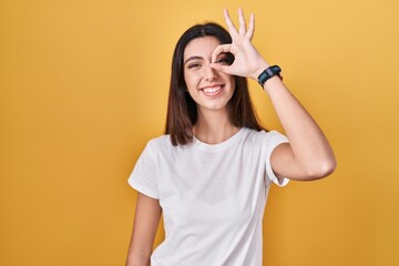 Young beautiful woman standing over yellow background doing ok gesture with hand smiling, eye looking through fingers with happy face.