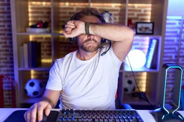 Sticker - Middle age man with beard playing video games wearing headphones covering eyes with arm, looking serious and sad. sightless, hiding and rejection concept