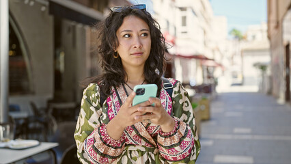 Wall Mural - Young beautiful hispanic woman using smartphone with serious expression at coffee shop terrace