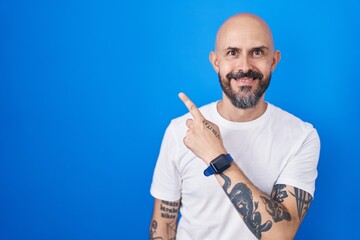 Poster - Hispanic man with tattoos standing over blue background cheerful with a smile on face pointing with hand and finger up to the side with happy and natural expression