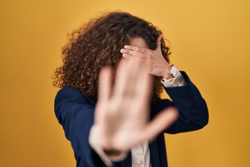 Sticker - Hispanic woman with curly hair standing over yellow background covering eyes with hands and doing stop gesture with sad and fear expression. embarrassed and negative concept.