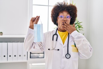 Poster - Young african american woman wearing doctor uniform holding safety mask covering mouth with hand, shocked and afraid for mistake. surprised expression