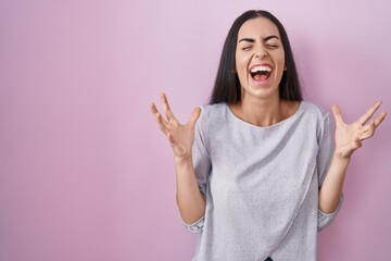 Wall Mural - Young brunette woman standing over pink background celebrating mad and crazy for success with arms raised and closed eyes screaming excited. winner concept