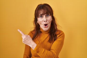 Wall Mural - Middle age hispanic woman standing over yellow background surprised pointing with finger to the side, open mouth amazed expression.