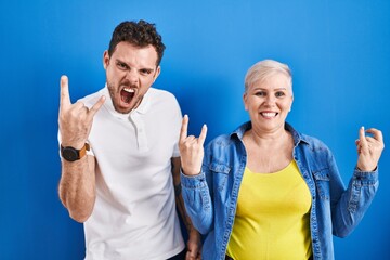 Wall Mural - Young brazilian mother and son standing over blue background shouting with crazy expression doing rock symbol with hands up. music star. heavy music concept.