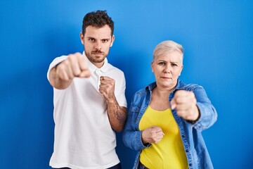 Poster - Young brazilian mother and son standing over blue background punching fist to fight, aggressive and angry attack, threat and violence