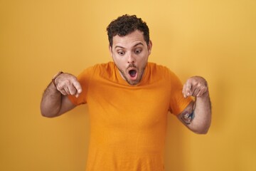 Sticker - Young hispanic man standing over yellow background pointing down with fingers showing advertisement, surprised face and open mouth