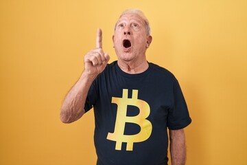 Sticker - Senior man with grey hair wearing bitcoin t shirt amazed and surprised looking up and pointing with fingers and raised arms.