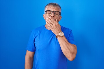 Wall Mural - Hispanic man with grey hair standing over blue background bored yawning tired covering mouth with hand. restless and sleepiness.