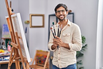 Young hispanic man artist smiling confident holding paintbrushes at art studio