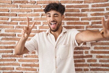 Poster - Arab man with beard standing over bricks wall background smiling with tongue out showing fingers of both hands doing victory sign. number two.