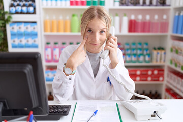 Canvas Print - Young caucasian woman working at pharmacy drugstore speaking on the telephone pointing to the eye watching you gesture, suspicious expression