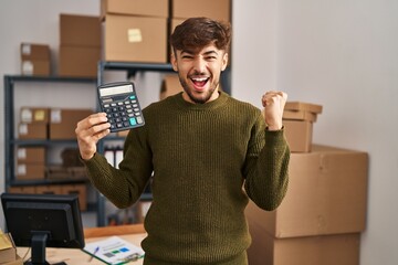 Canvas Print - Arab man with beard working at small business ecommerce holding calculator screaming proud, celebrating victory and success very excited with raised arms