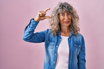 Wall Mural - Middle age woman standing over pink background smiling and confident gesturing with hand doing small size sign with fingers looking and the camera. measure concept.