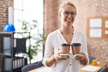 Poster - Young caucasian woman working at the office holding coffee cups winking looking at the camera with sexy expression, cheerful and happy face.