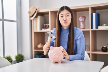 Canvas Print - Chinese young woman holding hammer and piggy bank puffing cheeks with funny face. mouth inflated with air, catching air.