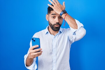 Wall Mural - Hispanic man with beard using smartphone typing message surprised with hand on head for mistake, remember error. forgot, bad memory concept.