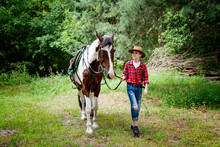 Little Handsome Blonde Smiling Boy In Red Checkered Shirt And Cowboy Brown Hat Walking And  Hugging Horse In Green Forest On Sunny Day	