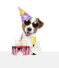 Poster - Jack russell terrier puppy wearing sunglasses and party hat holds birthday cake with lot of candle and glass of champagne above empty white board. isolated on white background