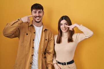 Wall Mural - Young hispanic couple standing over yellow background pointing with hand finger to face and nose, smiling cheerful. beauty concept