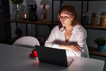 Wall Mural - Middle age hispanic woman using laptop at home at night making fish face with lips, crazy and comical gesture. funny expression.