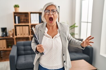 Sticker - Middle age woman with grey hair at consultation office angry and mad screaming frustrated and furious, shouting with anger. rage and aggressive concept.