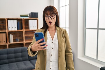 Wall Mural - Young brunette woman working at the office with smartphone scared and amazed with open mouth for surprise, disbelief face