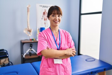 Canvas Print - Young brunette woman working at rehabilitation clinic happy face smiling with crossed arms looking at the camera. positive person.