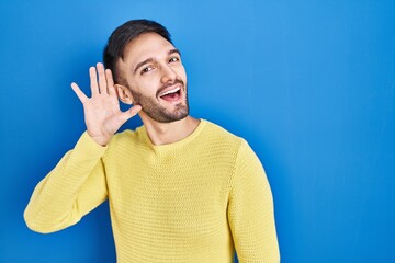 Canvas Print - Hispanic man standing over blue background smiling with hand over ear listening an hearing to rumor or gossip. deafness concept.