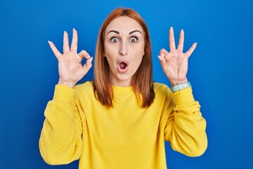 Canvas Print - Young woman standing over blue background looking surprised and shocked doing ok approval symbol with fingers. crazy expression