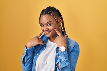 Sticker - African american woman with braids standing over yellow background smiling with open mouth, fingers pointing and forcing cheerful smile