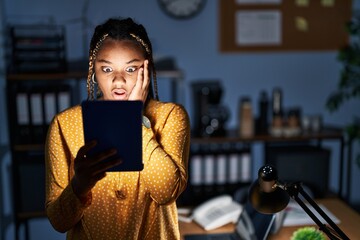 Wall Mural - African american woman with braids working at the office at night with tablet afraid and shocked, surprise and amazed expression with hands on face