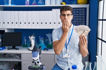 Canvas Print - Young hispanic man working at scientist laboratory holding dollars covering mouth with hand, shocked and afraid for mistake. surprised expression