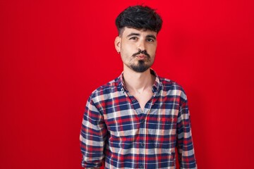 Wall Mural - Young hispanic man with beard standing over red background looking at the camera blowing a kiss on air being lovely and sexy. love expression.