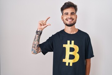 Sticker - Young hispanic man with tattoos wearing bitcoin t shirt smiling and confident gesturing with hand doing small size sign with fingers looking and the camera. measure concept.