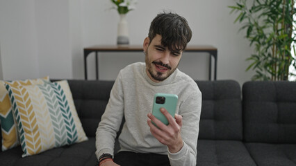 Poster - Young hispanic man using smartphone sitting on sofa at home