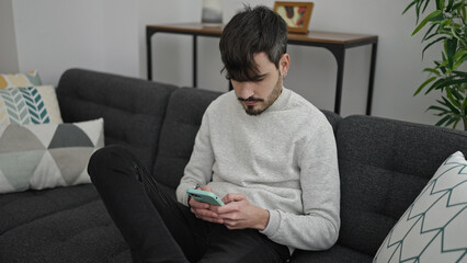 Wall Mural - Young hispanic man using smartphone sitting on the sofa at home