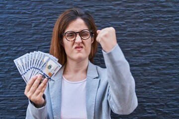 Wall Mural - Brunette woman holding dollars annoyed and frustrated shouting with anger, yelling crazy with anger and hand raised