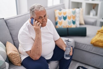 Wall Mural - Middle age grey-haired man talking on smartphone holding bottle of water at home