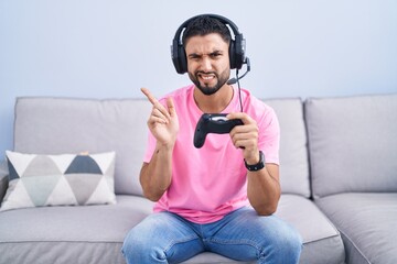 Canvas Print - Hispanic young man playing video game holding controller sitting on the sofa pointing aside worried and nervous with forefinger, concerned and surprised expression