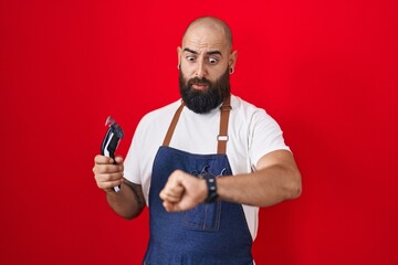 Wall Mural - Young hispanic man with beard and tattoos wearing barber apron holding razor looking at the watch time worried, afraid of getting late