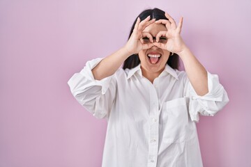 Sticker - Young brunette woman standing over pink background doing ok gesture like binoculars sticking tongue out, eyes looking through fingers. crazy expression.