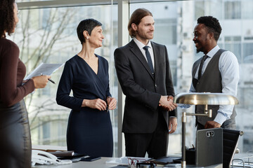 Multiethnic group of smiling business partners shaking hands in office standing against big windows, copy space