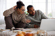 © Dmytro Hai - multiracial couple in warm clothes talking near laptop and books during online education at home on bed
