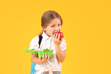 Poster - Happy little girl with backpack and lunchbox eating apple on yellow background