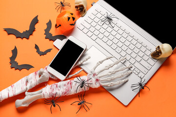 Skeleton hands with modern mobile phone, laptop and paper bats for Halloween celebration on orange background