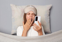Sleepy Woman In White T-shirt And Sleeping Eye Mask Lie In Bed On Pillow Under Blanket Isolated On Gray Background Using Phone Long Hours Being Tired Yawning